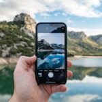 Person holding a smartphone and capturing a scenic mountain lake landscape, showcasing mobile camera photography — Photo by Unsplash